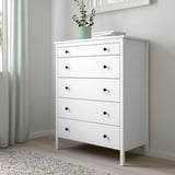 KOPPANG white chest with 5 drawers, black knobs, green plant, books, on rug, beside wall.
