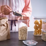 Person pours oats into transparent, airtight jar from paper bag. Jars stackable, with ventilation button for easy opening.