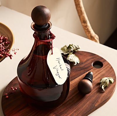 A brown glass bottle with a wooden stopper and label saying herbes vinager placed on a wooden tray.