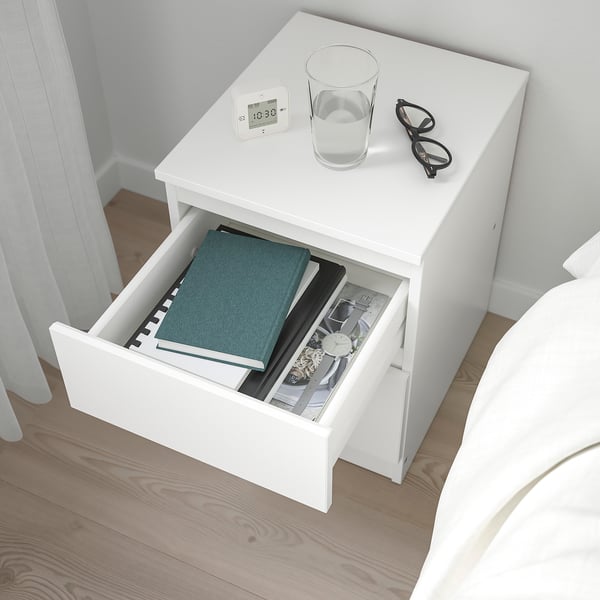 White bedside table with open drawer holding books, a glass of water, glasses, and a clock on top.