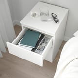 White bedside table with open drawer holding books, a glass of water, glasses, and a clock on top.