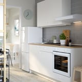 Modern kitchen with white cabinets, clock, oven, & sink. Green plant and dishes are on the counter.