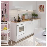 Modern white kitchen with wooden counter, stainless steel appliances, hanging utensils, plants, lemons, and colorful rug.