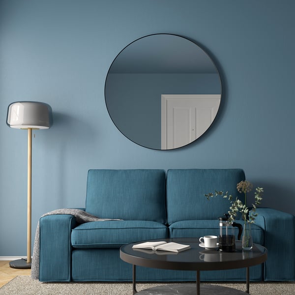 Modern living room with large round black-framed LINDBYN mirror above blue couch, complementing blue walls, and a grey floor lamp.