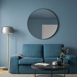 Modern living room with large round black-framed LINDBYN mirror above blue couch, complementing blue walls, and a grey floor lamp.