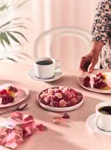 A cozy table setting with pink berry candies, white coffee cups, and plates of desert with raspberries.
