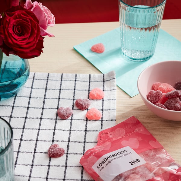Pink heart-shaped candies scattered on a checked cloth, with roses, a glass of water, and a candy bag labeled LÖRDAGSGODIS nearby.