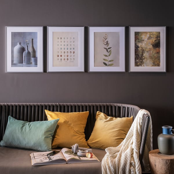 Four framed prints above a brown sofa with mustard pillows. Beside the sofa, a book, apple, blanket, and vases create a cozy space.