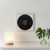 A framed black vinyl record hung on a light gray wall above a white shelf. The shelf holds a small lamp and a glass vase with green leaves.