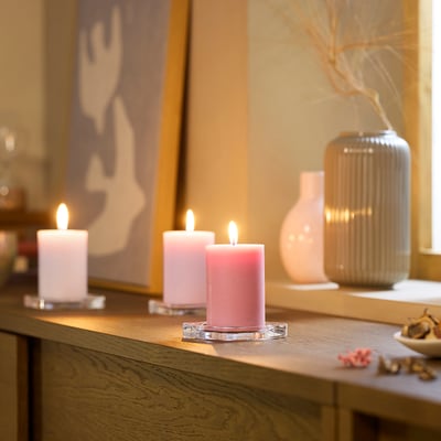 Three pink and white candles on wood. Pink ones in holders, plant beside. Abstract art in background. Cosy, scented.