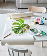 A craft table set with MÅLA paints, a large green leaf, brushes, and paper roll. Minimalist design with white and wood tones.