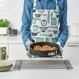 Person wearing apron holds springform pan with baked rolls. Apron has kitchen tool prints. Pan sits on cooling rack with stacked plates and cups.