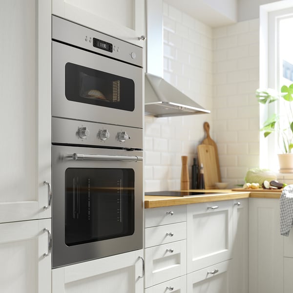 Modern kitchen with built-in stainless steel oven and microwave, digital displays, silver handles, and wooden countertop with utensils and plants.
