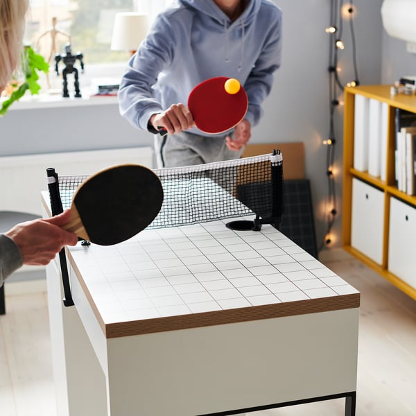 Two people play table tennis with a red and black paddle on a white table with a net in a room with a robot toy and books.