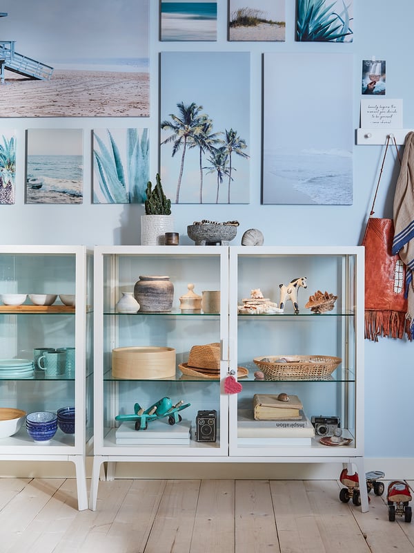 White MILSBO display cabinet with glass doors, holding various decorative items, against a wall with beach-themed artwork.