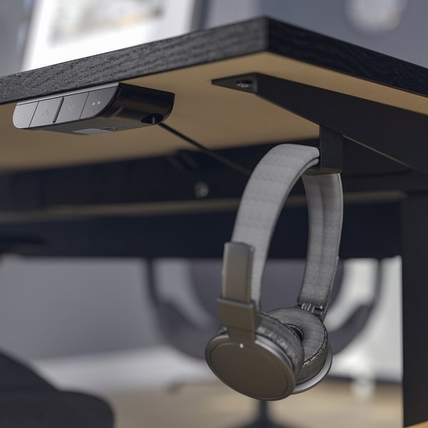 Wireless headphones hang on hook beneath modern desks sleek surface and cable management tray.