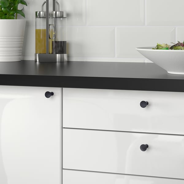 A modern kitchen with white cabinets and black countertops black NYDALA knobs, enhancing cabinet style. A salad bowl is on the sleek countertop.