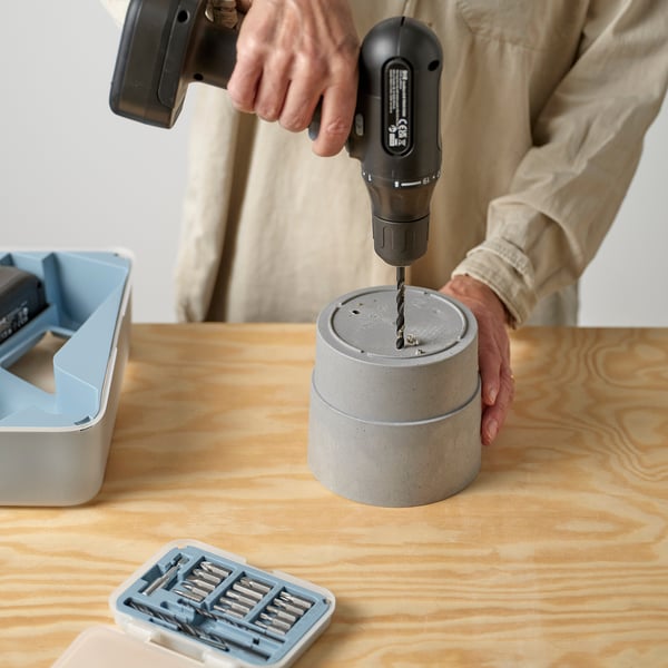 Person drilling a hole in a concrete-like planter on a wooden table, using a power drill; plastic drill bit holder and container nearby.