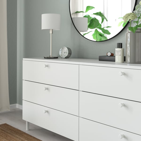 White dresser with six drawers, each having round knobs for easy gripping. Topped with a round mirror, lamp, and decorative items.