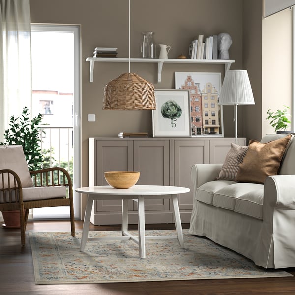 Modern living room with colorful rug, grey armchair, white coffee table, wooden side table, wicker lamp, and decorations.