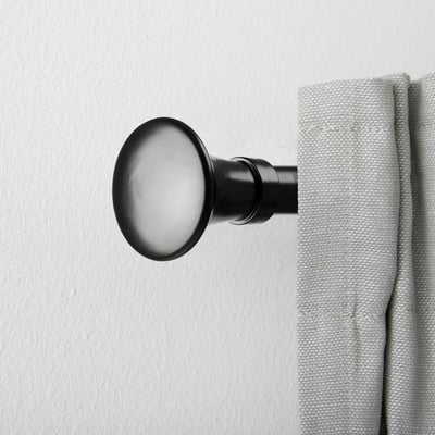 Black curtain rod finial with trumpet shape, holding light gray curtain against white wall.