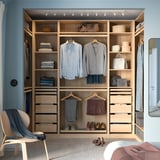 Wooden closet with shelves, drawers, and lighting. Neatly organized clothes, chair with blanket, glazed door.
