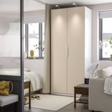 Modern bedroom with a beige PAX wardrobe between windows, comfortable chair, side table with books, bed in background.