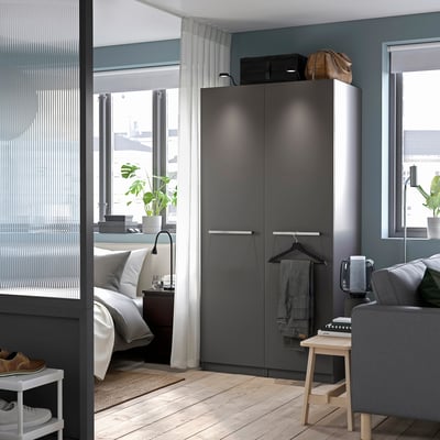 Modern apartment bedroom with dark gray PAX wardrobe, white bed, large windows, and wooden floor.