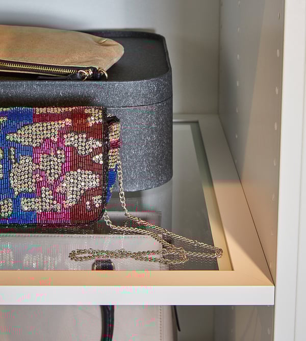 A white PAX wardrobe with clear shelf, holding a black hatbox, and a colorful beaded handbag with a gold chain strap.