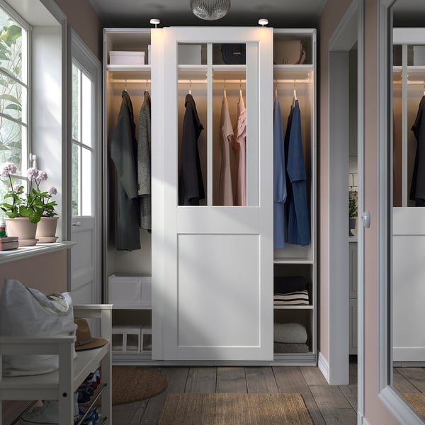 Modern closet with white PAX wardrobes, glass doors, and mirrors. Soft lighting, wood floor, plants, bench.