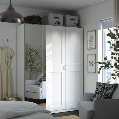 A white PAX wardrobe with two doors, a mirror, and storage boxes, positioned beside a window with green plants and framed pictures.