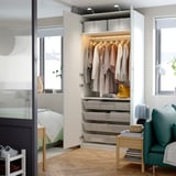 Open wardrobe displaying clothes, with shelves and drawers below, in modern bedroom next to green chair and window.