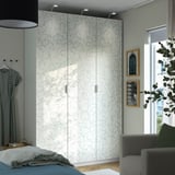 Modern bedroom with large white floral-patterned PAX wardrobe, grey chair, and large window. Wardrobe stands tall between other furniture.