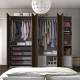 A well-organized, open wardrobe displaying hangers with clothes, shelves, and storage boxes. White lamp above, bed in front.