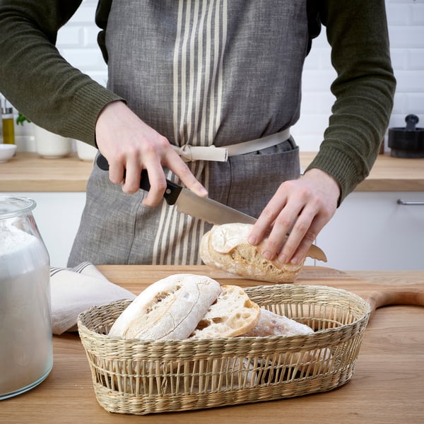 A person slices bread using a knife. Bread pieces in a woven basket on a table. A jar and cloth are also visible.