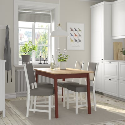Small, modern dining set with wooden table and gray cushioned chairs, positioned neatly under a window, between white kitchen cabinets.