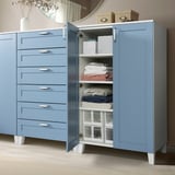 Light blue storage unit with drawers and open cabinet showing folded towels and boxes, on light brown floor.