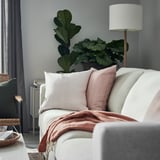 Modern living room with white sofa, pink cushions, red blanket, tall lamp, and large green plants.