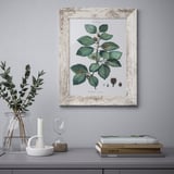 Framed botanical print above table with greenery, candle, books.