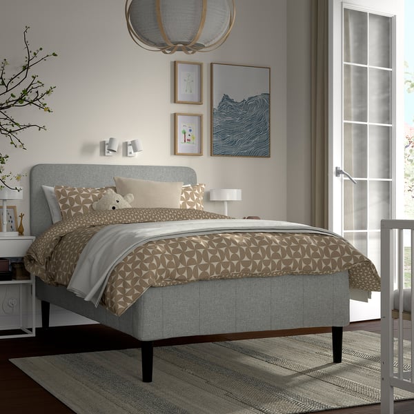 Modern bedroom with grey RÅHOLMAR bed, patterned duvet, nightstands, artwork, ceiling light, and rug by crib.