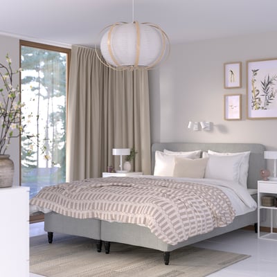Modern, neutral bedroom with light grey bed, patterned bedding, white pillows, beige curtains, hanging light, small nightstands, and decorated wall.