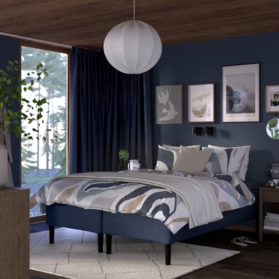 Modern bedroom with dark blue walls, large bed under a pendant lamp, patterned bedding, wood furniture, and green plants.