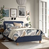 Bedroom with blue RÅHOLMAR bed, abstract bedding, wooden legs, large rug, wall art.