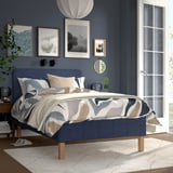 Modern blue bed with abstract print bedding, aligned wall art, and large photo window. Light wood nightstand and floor lamp.
