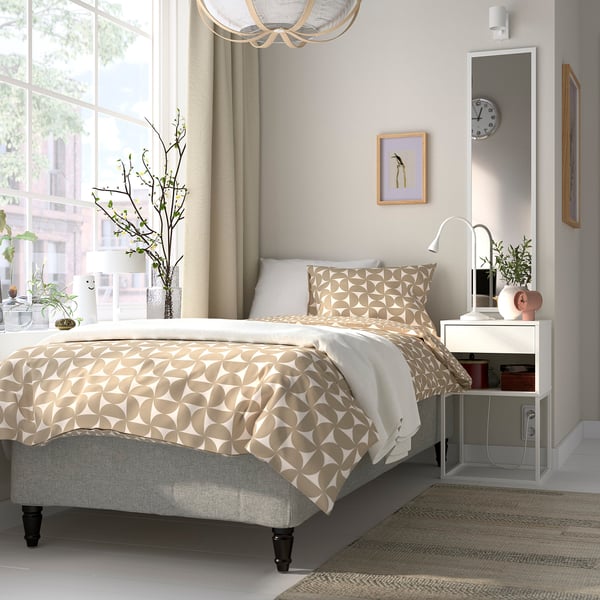 Modern neutral-toned bedroom with RÅHOLMAR bed. Bed has patterned beige and white bedding, nightstands with decor, and large windows with curtains.