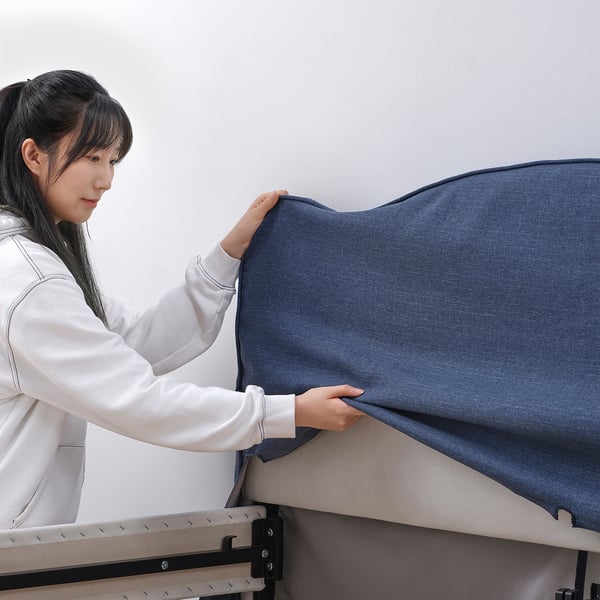 A woman is putting on a blue cover on a metal bed frame, showcasing its easy-to-remove and machine-washable design.