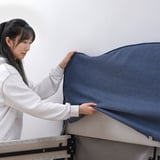 A woman is putting on a blue cover on a metal bed frame, showcasing its easy-to-remove and machine-washable design.
