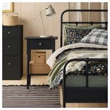 Bedroom with metal bed, dark green blanket, floral pillow, black nightstand, lamp, and striped rug.