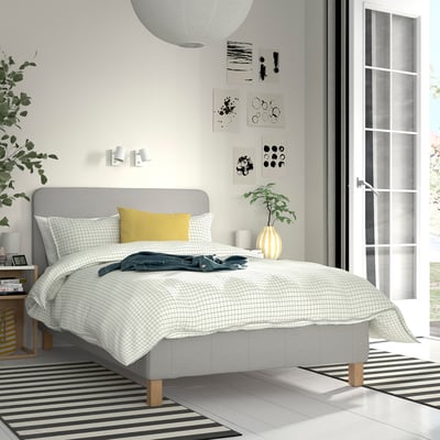 Modern bedroom with grey bed, white linens, yellow pillow, striped rug, plants, and wall art. Spacious with glass door.