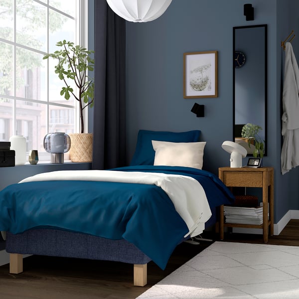 Modern blue RENFJÄLLET bed in cozy room. Blue/white bedding, wooden nightstand with lamp & plant. Large window, plant beside bed.